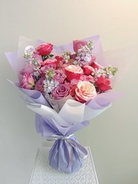 A bouquet of pink, white, and purple flowers including roses, lilies, and orchids wrapped in lavender paper.