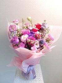 A bouquet of pink, white, and purple flowers including roses, lilies, and orchids, wrapped in soft pastel paper.