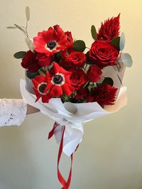 A bouquet of vibrant red roses and anemones with lush greenery, wrapped in white paper and a red ribbon