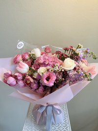 Pink, white, and purple floral bouquet featuring roses, lilies, and orchids wrapped in pastel paper with a gray ribbon