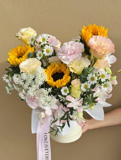 A cheerful vase bouquet with sunflowers, carnations, lisianthuses, daisies, and orchids, tied with a pink ribbon that reads “Love is Eternal.”