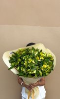 Person holding a large hand-tied bouquet of yellow-green Alstroemeria flowers with abundant foliage, wrapped in cream paper with yellow ribbon