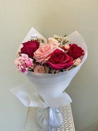 Bouquet of pink and red roses with pink carnations and baby's breath wrapped in white paper.