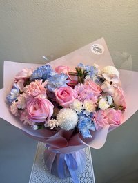A bouquet of pink roses, white lilies, and purple orchids wrapped in pastel paper with a purple ribbon.